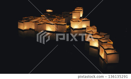 People sending glowing paper lantern boats onto a dark reflective river during a night festival. People sending glowing paper lantern boats onto a dark reflective river during a night festival. 136135291
