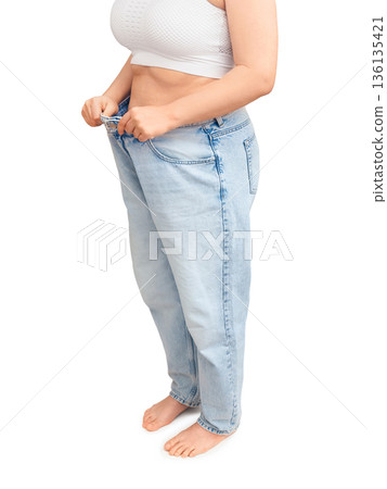 Slim woman showing weight loss result in oversized jeans. Fit female pulling loose pants away from waist isolated on white background. Diet success concept Slim woman showing weight loss result in oversized jeans. Fit female pulling loose pants away from waist isolated on white background. Diet success concept 136135421