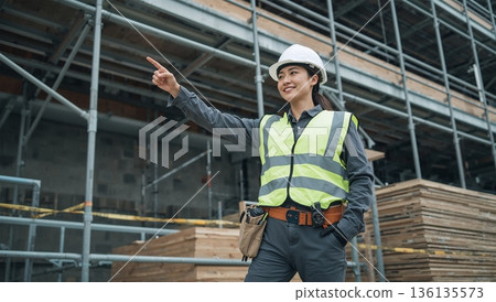 在日本一處建築工地，一名頭戴安全帽、身穿安全背心的女子站在鷹架前指著前方。 136135573