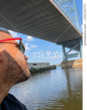 A man wearing sunglasses is looking at Benjamin Franklin bridge over a body of water A man wearing sunglasses is looking at Benjamin Franklin bridge over a body of water 136135773