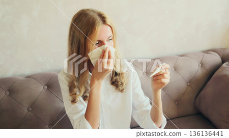 Sick young woman sneezing blow nose using tissue and measuring checking body temperature at home 136136441