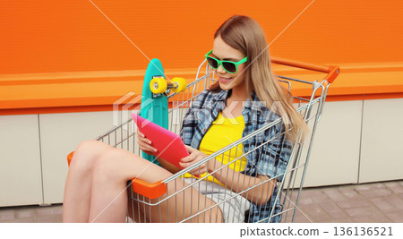Modern young woman using digital tablet computer sitting in shopping trolley cart on city street 136136521