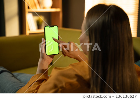 Woman relaxing on a couch at home while holding a modern smartphone with a bright green screen, ideal for overlaying custom content, advertising, or app interfaces Woman relaxing on a couch at home while holding a modern smartphone with a bright green screen, ideal for overlaying custom content, advertising, or app interfaces 136136627