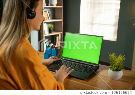 Young woman in her 20s wearing a headset and smiling while typing on a laptop with a green screen, working remotely from a bright home office and joining a virtual meeting Young woman in her 20s wearing a headset and smiling while typing on a laptop with a green screen, working remotely from a bright home office and joining a virtual meeting 136136634
