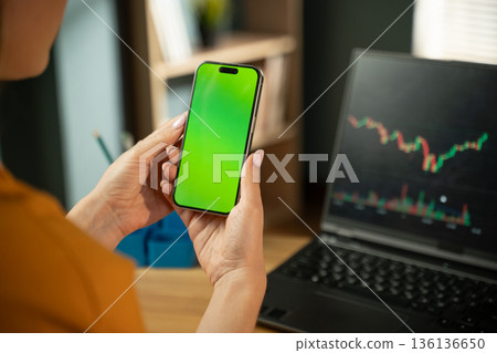 Person holding a smartphone with a green screen, representing digital trading and financial analytics, with a laptop displaying stock market charts in the background Person holding a smartphone with a green screen, representing digital trading and financial analytics, with a laptop displaying stock market charts in the background 136136650
