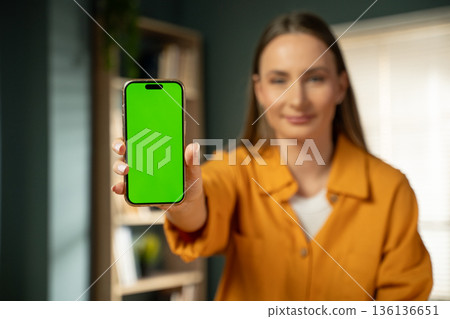 Young adult woman holding a vertical smartphone screen forward, displaying a customizable green screen for mobile app or content promotion, with her smiling face blurred in the background Young adult woman holding a vertical smartphone screen forward, displaying a customizable green screen for mobile app or content promotion, with her smiling face blurred in the background 136136651