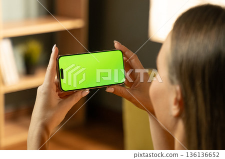 Woman holding a mobile phone horizontally in her hands, viewing a green screen display, providing a customizable mockup for various digital content like apps or videos Woman holding a mobile phone horizontally in her hands, viewing a green screen display, providing a customizable mockup for various digital content like apps or videos 136136652