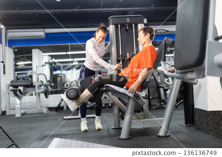 Personal trainer guiding senior woman on leg machine 136136749