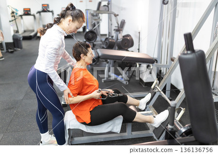 Personal trainer assisting senior woman exercising using gym machine 136136756
