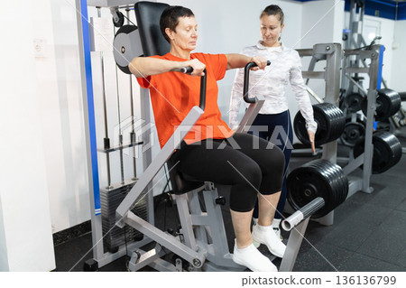 Senior woman training on gym machine with personal trainer 136136799