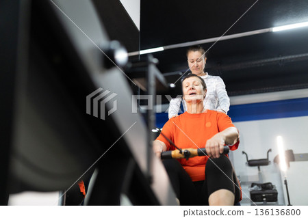 Senior woman training on rowing machine with personal trainer 136136800