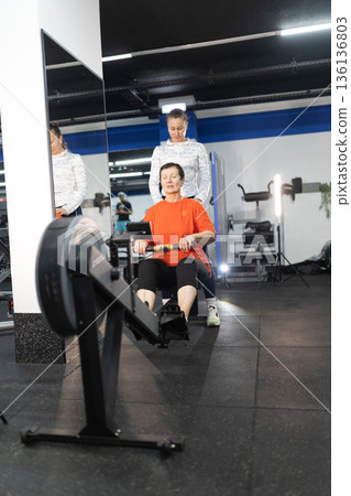 Senior woman receiving personal training on rowing machine 136136803