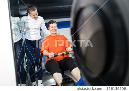 Senior woman training rowing machine with personal trainer 136136806
