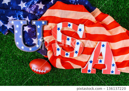 USA letters rest on flag with football nearby on grass, festive sports theme American pride. 136137220