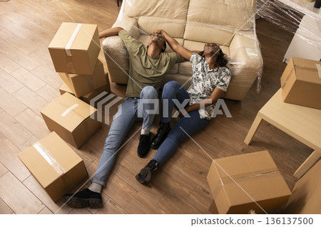 Black couple with heads on sofa, resting after hard moving day. African american young homeowners bonding and enjoying relocation, sitting on floor with packed cardboard boxes in new home. 136137500