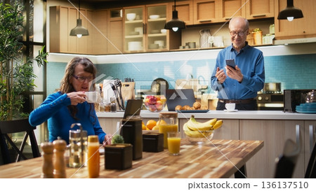 Senior adult browsing internet on smartphone and smiles in kitchen, relaxing with coffee and enjoying peaceful retired life. Retirement wellbeing, positive lifestyle and routine. Camera A. 136137510