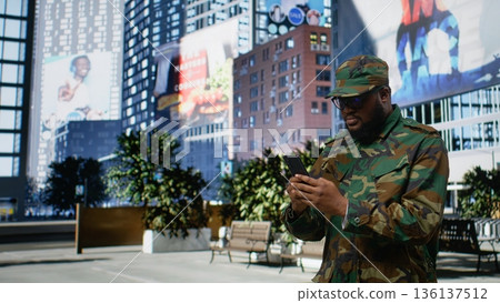 African american service person in full gear opens mobile phone map, walking through a modern downtown area in uniform. Soldier admiring skyscrapers and busy city street life. Camera A. 136137512