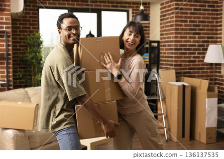 Joyful multiethnic spouses standing indoors holding cardboard box on moving day. Happy african american man and caucasian woman posing in modern apartment, celebrating home ownership together. Joyful multiethnic spouses standing indoors holding cardboard box on moving day. Happy african american man and caucasian woman posing in modern apartment, celebrating home ownership together. 136137535