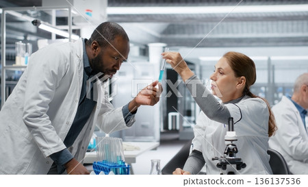 Teamworking lab researchers inspect liquids in glass flasks during clinical trials. Team of research facility colleagues comparing chemicals in glassware containers, testing formula, camera B 136137536