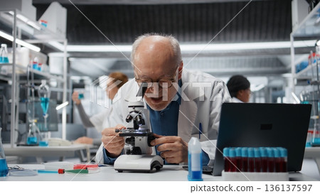 Pathology technician analyzing cellular structures using professional medical microscope. Elderly man conducting microscopic sample examination in sterile clinical lab environment, camera A Pathology technician analyzing cellular structures using professional medical microscope. Elderly man conducting microscopic sample examination in sterile clinical lab environment, camera A 136137597