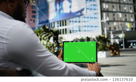 Horizontal green screen with black man sitting on a downtown bench, holding tablet with mockup and enjoying metropolitan area vibes. Contemporary activity under sunlight, city life. Camera A. 136137610