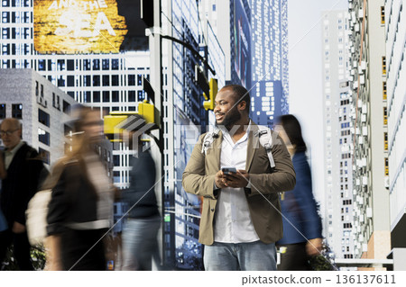 Street portrait of black adult with backpack using mobile phone and walking through the city surrounded by pedestrians, highlighting modern activity, routine and vibrant urban street life. 136137611