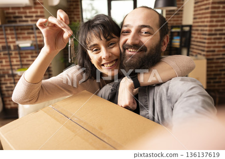 Excited young couple posing with house keys after moving into new brick wall apartment. Smiling caucasian man and woman hugging, holding cardboard box, celebrating first home together. 136137619