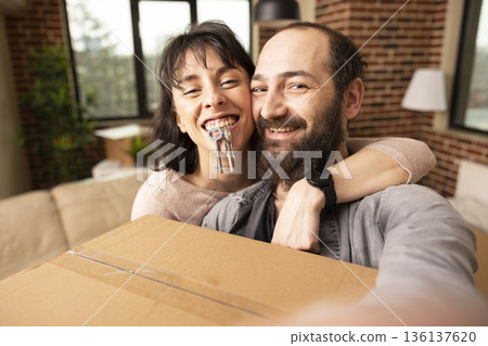 Happy couple taking selfie in new home as white woman bites house keys and hugs boyfriend. Caucasian man holds box and captures fun moment with wife in brick wall apartment on moving day. 136137620