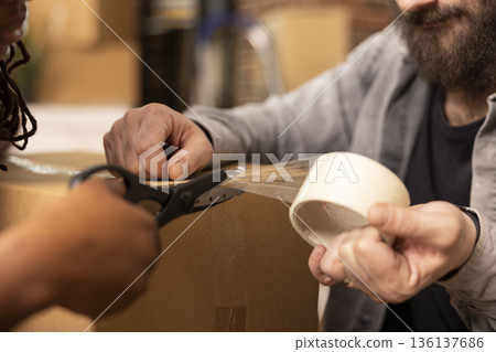 Closeup of black woman cutting adhesive tape as caucasian man seals cardboard box for moving. Scissors and tape visible during multiethnic couple packing process, preparing belongings for relocation. 136137686