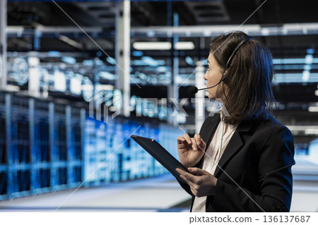 Data center customer support agent helping users, chatting with them using headset mic. Woman in server room talking in microphone, providing remote tech assistance to business clients 136137687