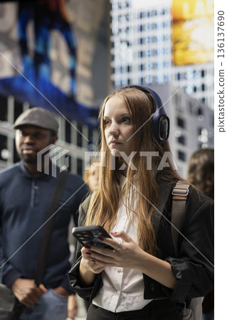 Gen z university student walking and browsing mobile apps on phone, wearing big headphones and a backpack. Young adult stylish cool girl waiting at a red light surrounded by pedestrians. 136137690