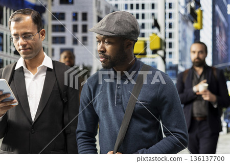 Portrait of casual african american man walking along the streets in the city, blurred motion of people behind reflecting realistic routine and urban street activity. Multicultural crowd. Portrait of casual african american man walking along the streets in the city, blurred motion of people behind reflecting realistic routine and urban street activity. Multicultural crowd. 136137700