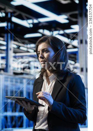 Technician in high tech data center using tablet, managing server virtualization. Programmer using touchscreen device in server hub, doing upgrades sustaining long term equipment rigs reliability 136137704