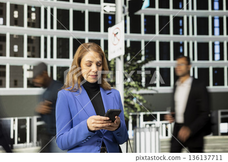 Portrait of active businesswoman professional browsing apps on busy city avenue, surrounded by a multicultural crowd in motion and highlighting urban lifestyle. Realistic everyday city life. 136137711
