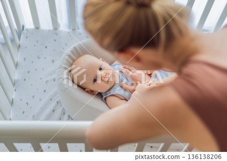 Mother gently sitting beside the crib and stroking her baby resting in a cozy cocoon nest. Tender intimate moment of care and trust at home. Motherhood, infant sleep and emotional bonding concept 136138206