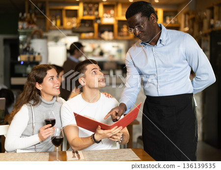 African waiter tells restaurant customers about the range of dishes and wines 136139535