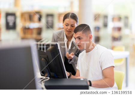 Male student works on computer in library and talks with female friend. 136139558