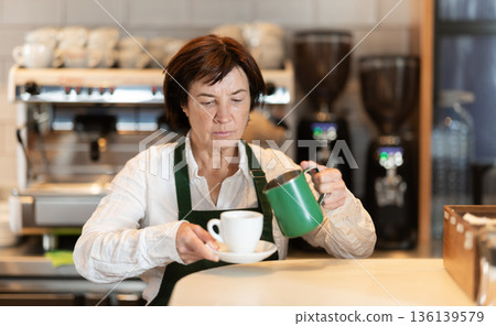 Senior woman cafe worker pours coffee from cezve coffee pot into cup Senior woman cafe worker pours coffee from cezve coffee pot into cup 136139579