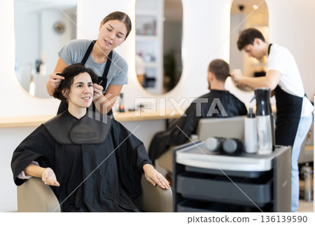 Young woman barbershop employee stands next to female client, discussing details of haircut 136139590