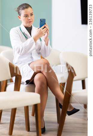 Attentive positive young girl medical student sitting and listening lecture, taking photo or QR with mobil in lecture hall Attentive positive young girl medical student sitting and listening lecture, taking photo or QR with mobil in lecture hall 136139733