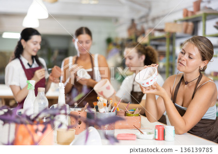 Young woman sits near table and decorates clay ceramic craft vase, plate Young woman sits near table and decorates clay ceramic craft vase, plate 136139785