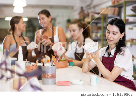 Young woman sits near table and decorates clay ceramic craft vase, plate 136139802