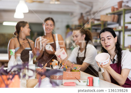 Young woman sits near table and decorates clay ceramic craft vase, plate Young woman sits near table and decorates clay ceramic craft vase, plate 136139826