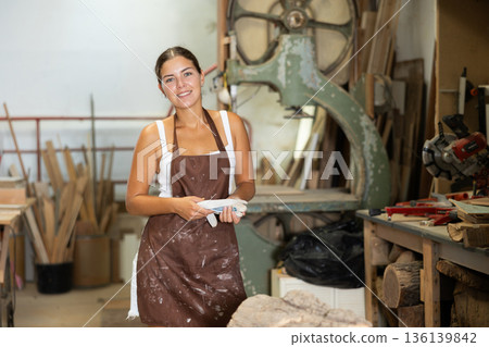 Girl employee of workshop cuts wooden board into parts using saw Girl employee of workshop cuts wooden board into parts using saw 136139842