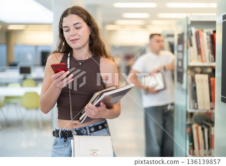 Female student communicates on mobile phone in the interior of public library Female student communicates on mobile phone in the interior of public library 136139857
