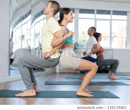 Group of men and women doing acroyoga exercises in pairs, exercising partner yoga during training in fitness center Group of men and women doing acroyoga exercises in pairs, exercising partner yoga during training in fitness center 136139861