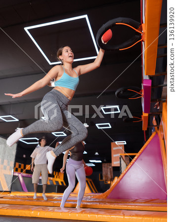 Excited young woman enjoying amusement activities while jumping high on trampoline and hitting basketball hoop with ball in game club 136139920