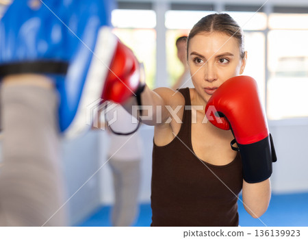 Woman wearing boxing gloves exercise and punch to pads for boxing. Woman with trainer at gym Woman wearing boxing gloves exercise and punch to pads for boxing. Woman with trainer at gym 136139923