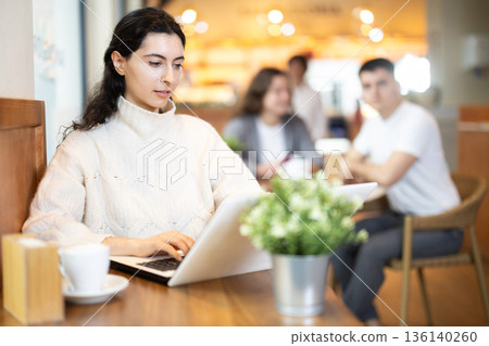 In cozy cafe hall, freelance girl works with laptop and drinks coffee. 136140260