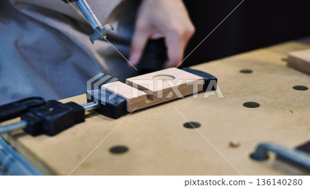 Woodworker drilling a circular hole into a wooden piece clamped on a workbench 136140280